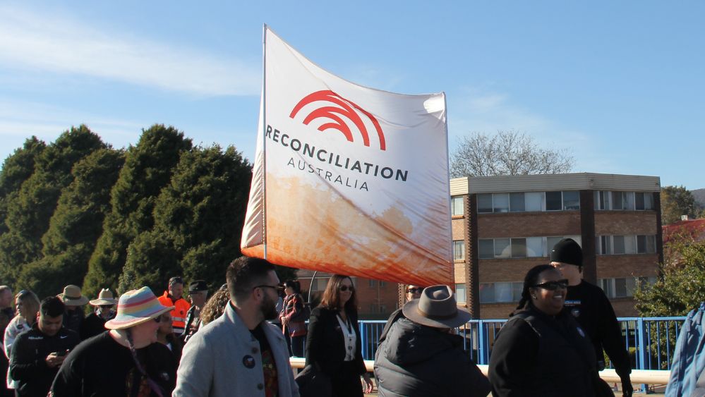 Reconciliation Australia staff carrying the banner amongst a large group of people walking together on a beautiful sunny day. This photo is from the Queanbeyan Reconciliation Walk 2025.