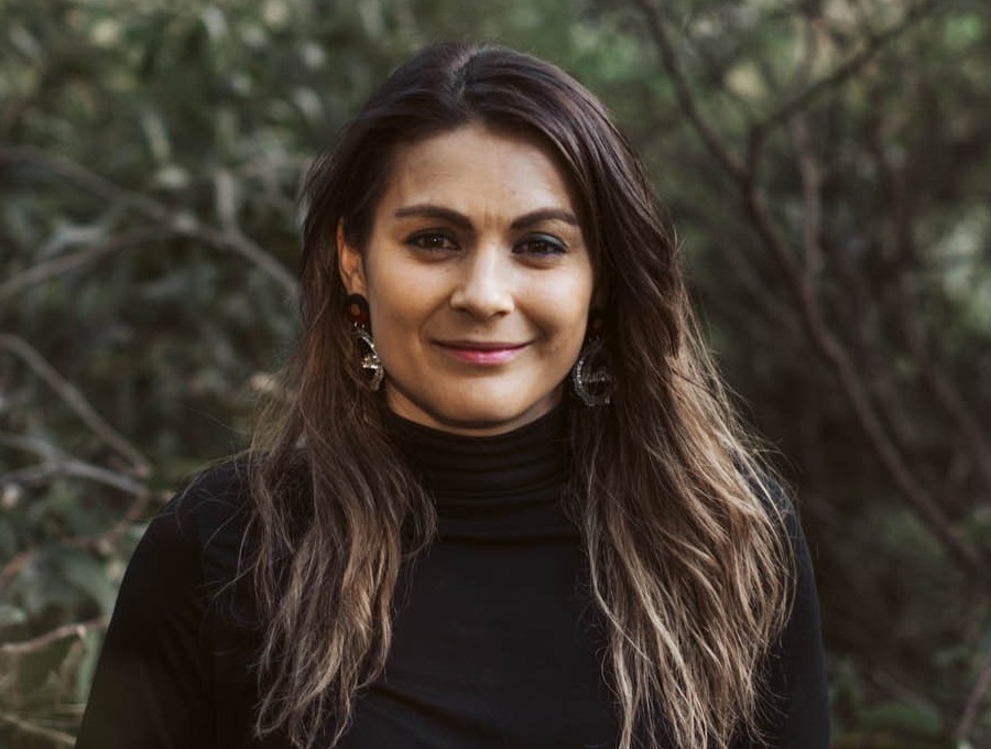 Muthi Muthi/Gunditjmara woman Jacara Egan, standing in the bush, smiling.