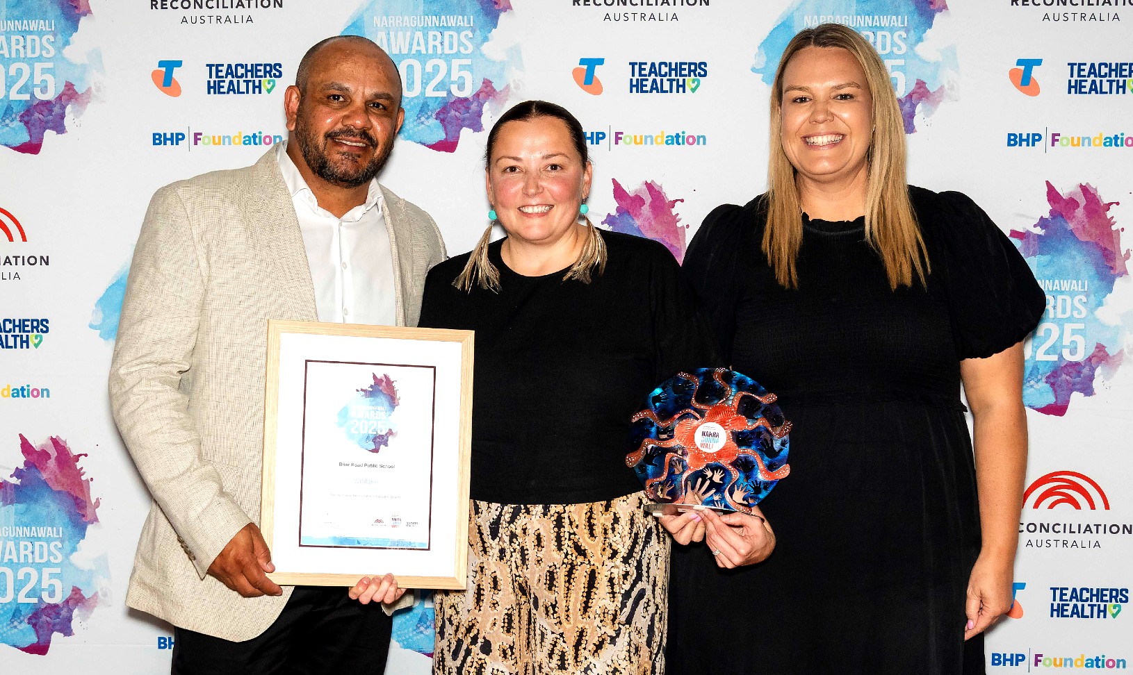 One man and two women holding a framed certificate and a crved metal trophy in front of a media banner.