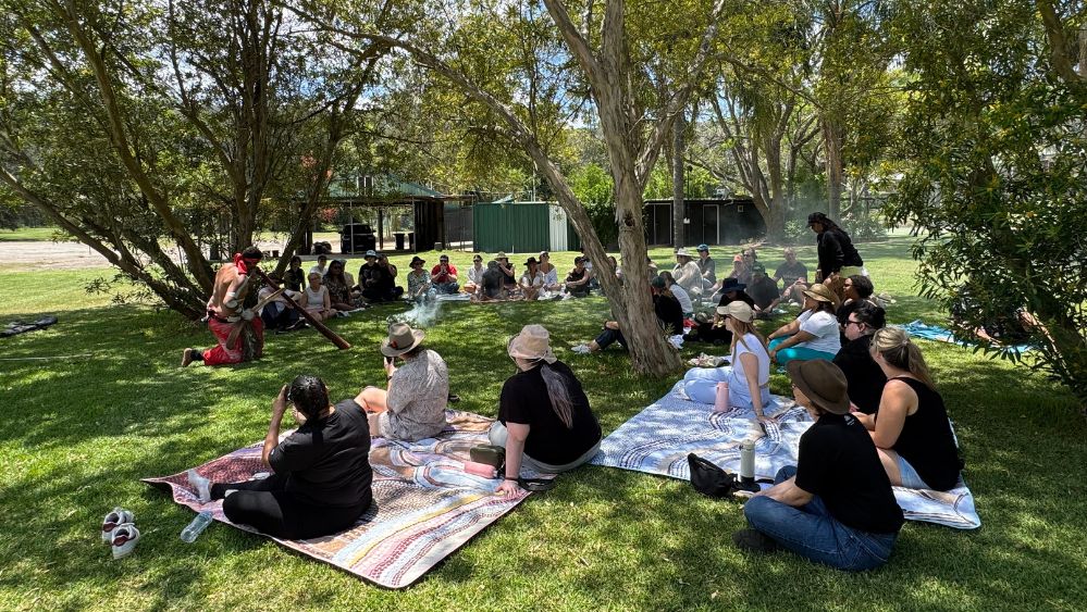 A group of people sitting in a loose circle on the grass, watching a man play didgeridoo as part of a smoking ceremony.