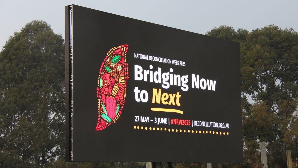 The National Reconciliation Week artwork and theme - Bridging Now to Next - on a digital billboard in a park in Melbourne.