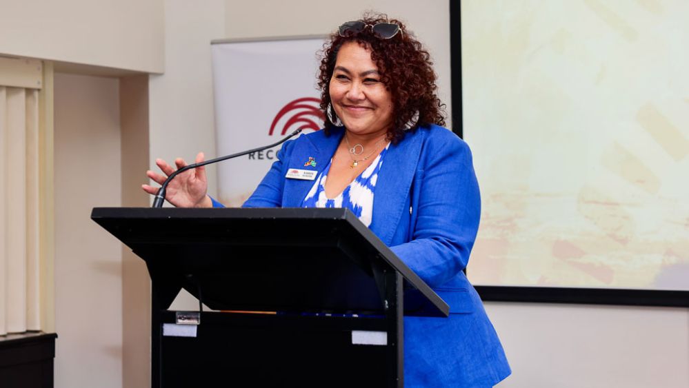 A middle-aged Aboriginal woman - Reconciliation Australia CEO Karen Mundine - standing at a lectern, smiling.