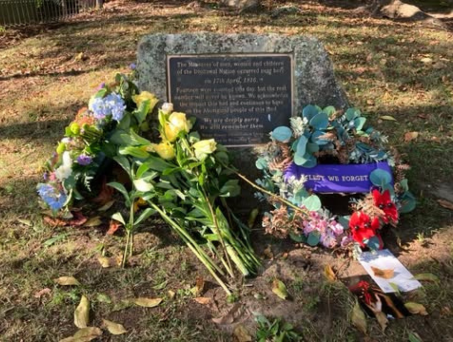 A photo of a plaque surrounded by flowers and wreaths. The plaque reads: 'The Massacre of men, women and children of the Dharawal Nation occurred near hre on 17th April 1816. Fourteen were counted this day, but the real number will never be known. We acknowledge the impact this had and continues to have on the Aboriginal people of this land. We are deeply sorry. We will remember them.'