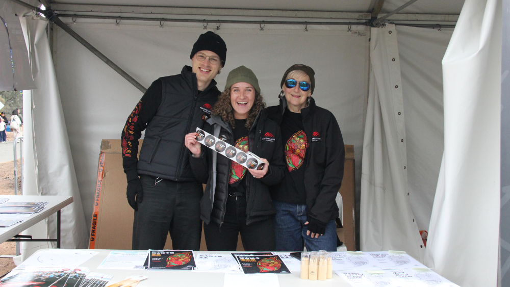 Three people - a man and two women - stand in a festival stall, wearing Reconciliation Australia and National Reconciliation Week 2025-branded clothing, showing off National Reconciliation Week 2025 merchandise. They are all smiling and look very happy.
