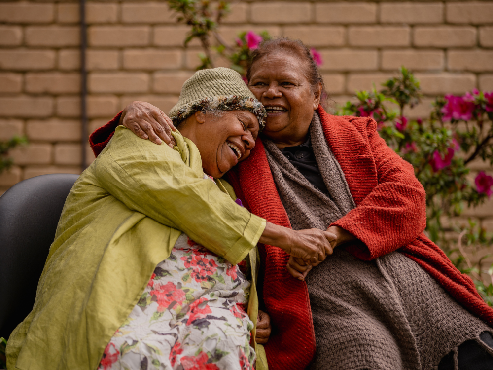 Two older woman embrace warmly, smiling.