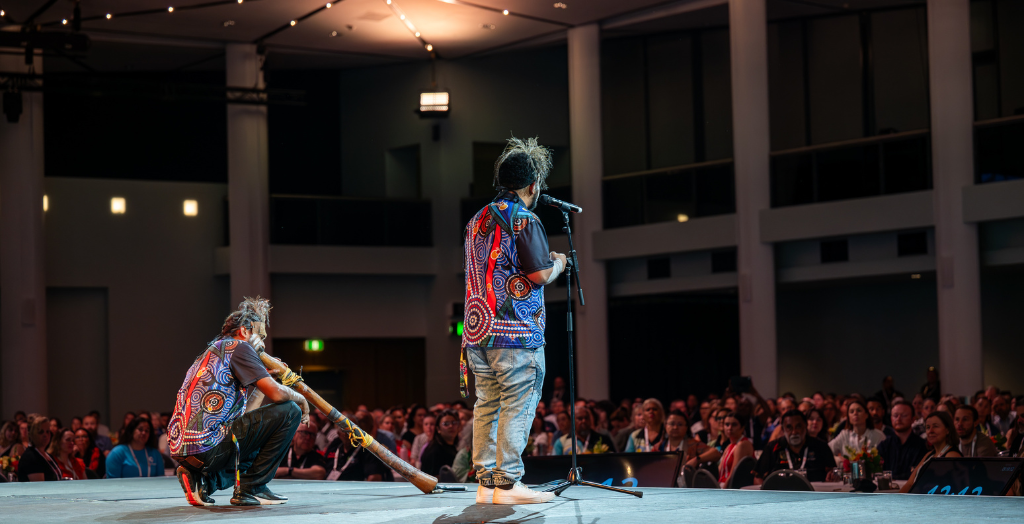 Two men seen from behind, on a stage in front a large group of people, seated. One of the men is kneeling and playing a didgeridoo-like instrument. The other man is standing at speaking into a microphone.