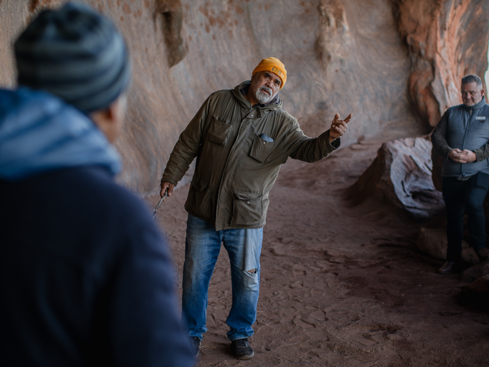 A man leading a tour stands in a shallow cave on red sand, pointing at something off camera.